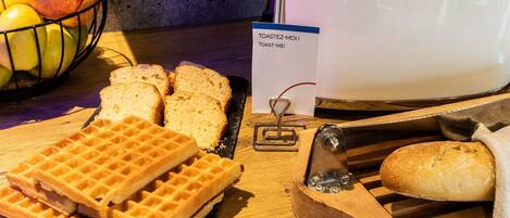 A kitchen counter with a toaster, a waffle maker, sliced bread, and a bowl of fruit.