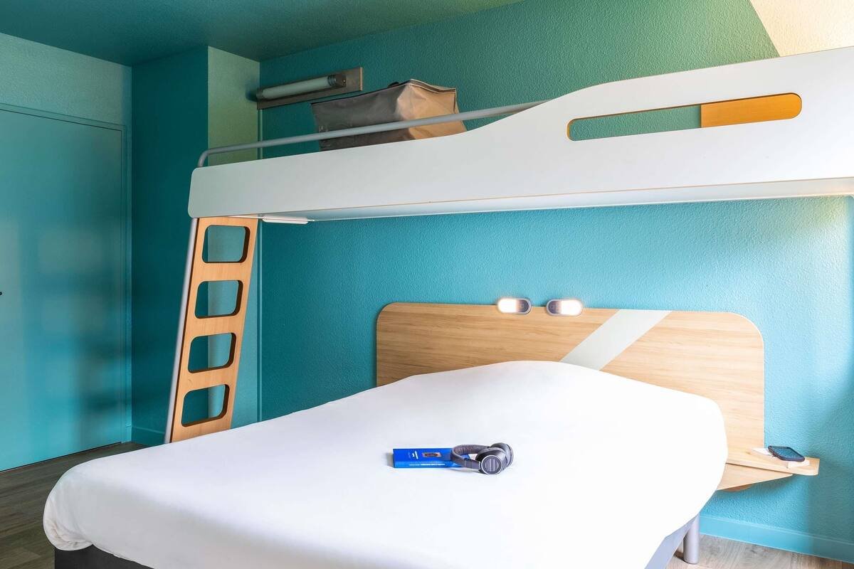 A bunk bed with a ladder, a bedside table with a book and headphones, and a bed with a white sheet.