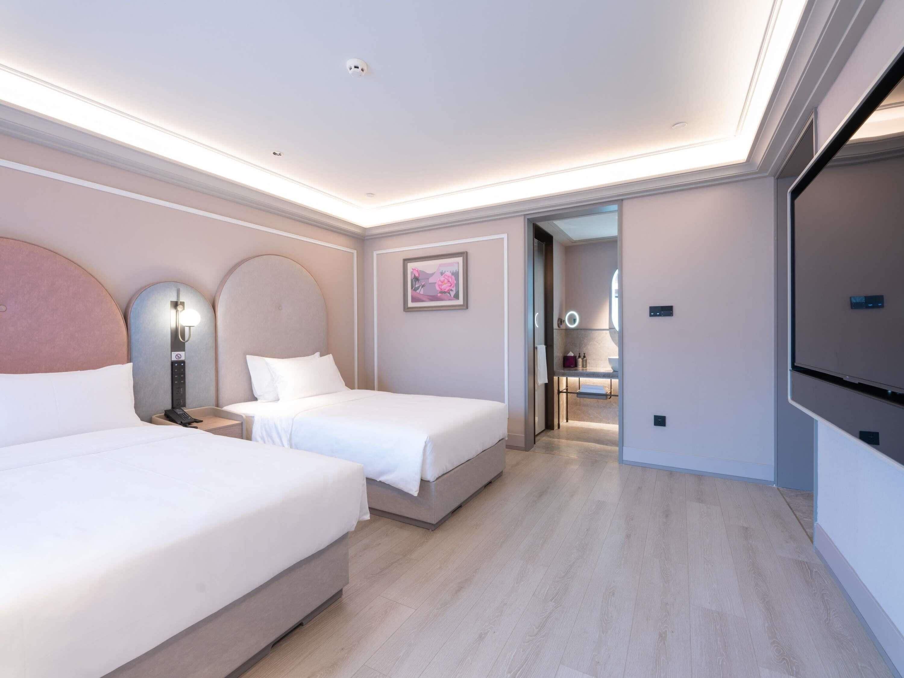 A modern hotel room with two beds, a large mirror, a vanity with a ring light, and a view into the bathroom.