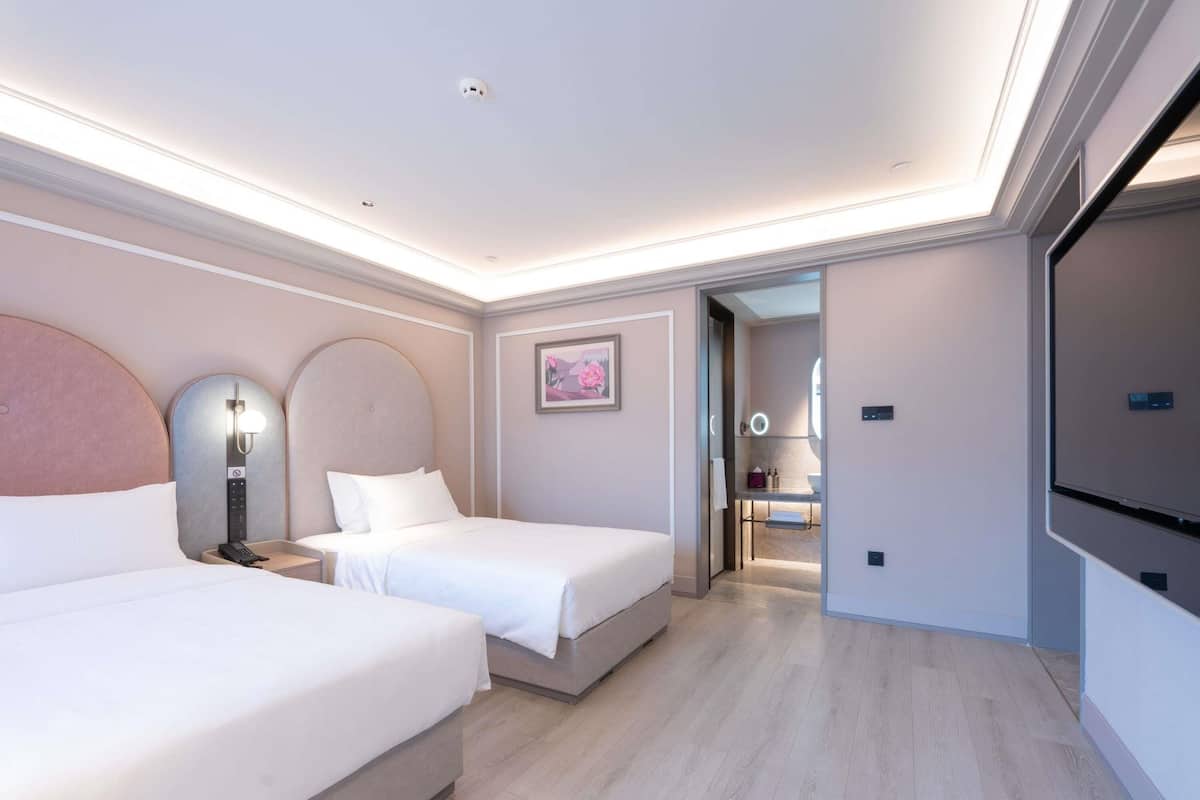 A modern hotel room with two beds, a large mirror, a vanity with a ring light, and a view into the bathroom.