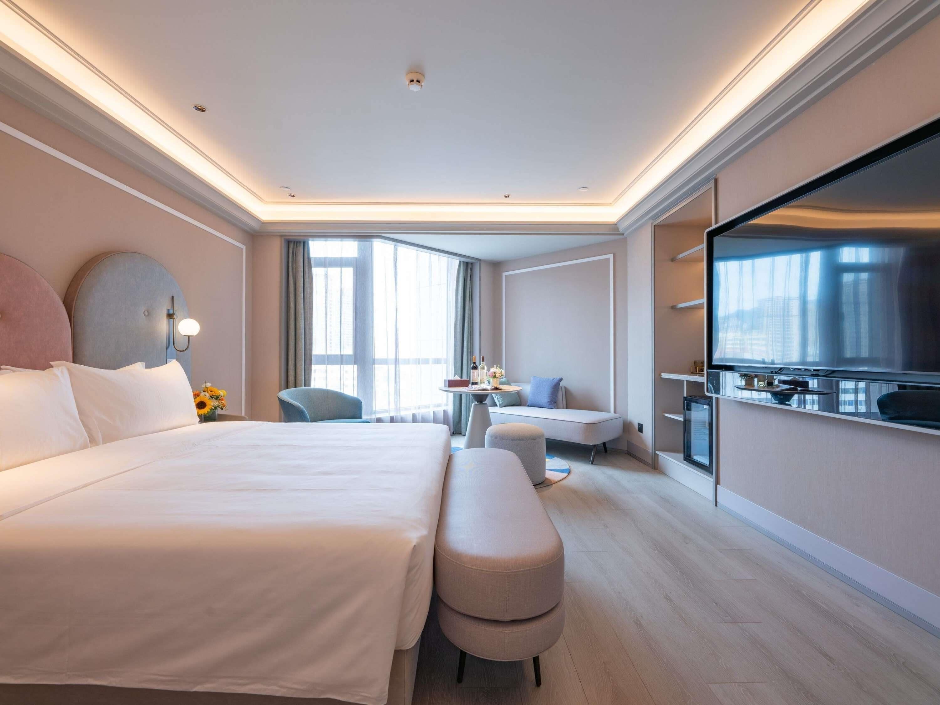 A modern hotel room with a large bed, a seating area with a table, a TV, and large windows.