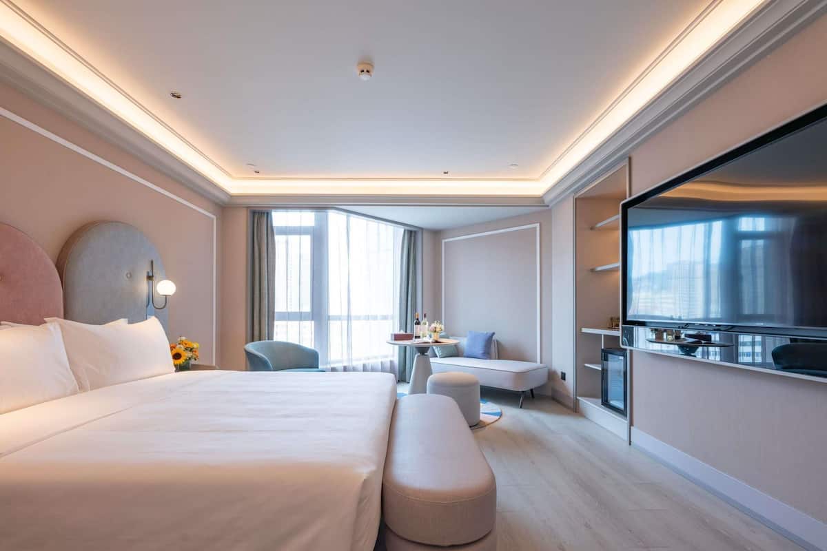 A modern hotel room with a large bed, a seating area with a table, a TV, and large windows.