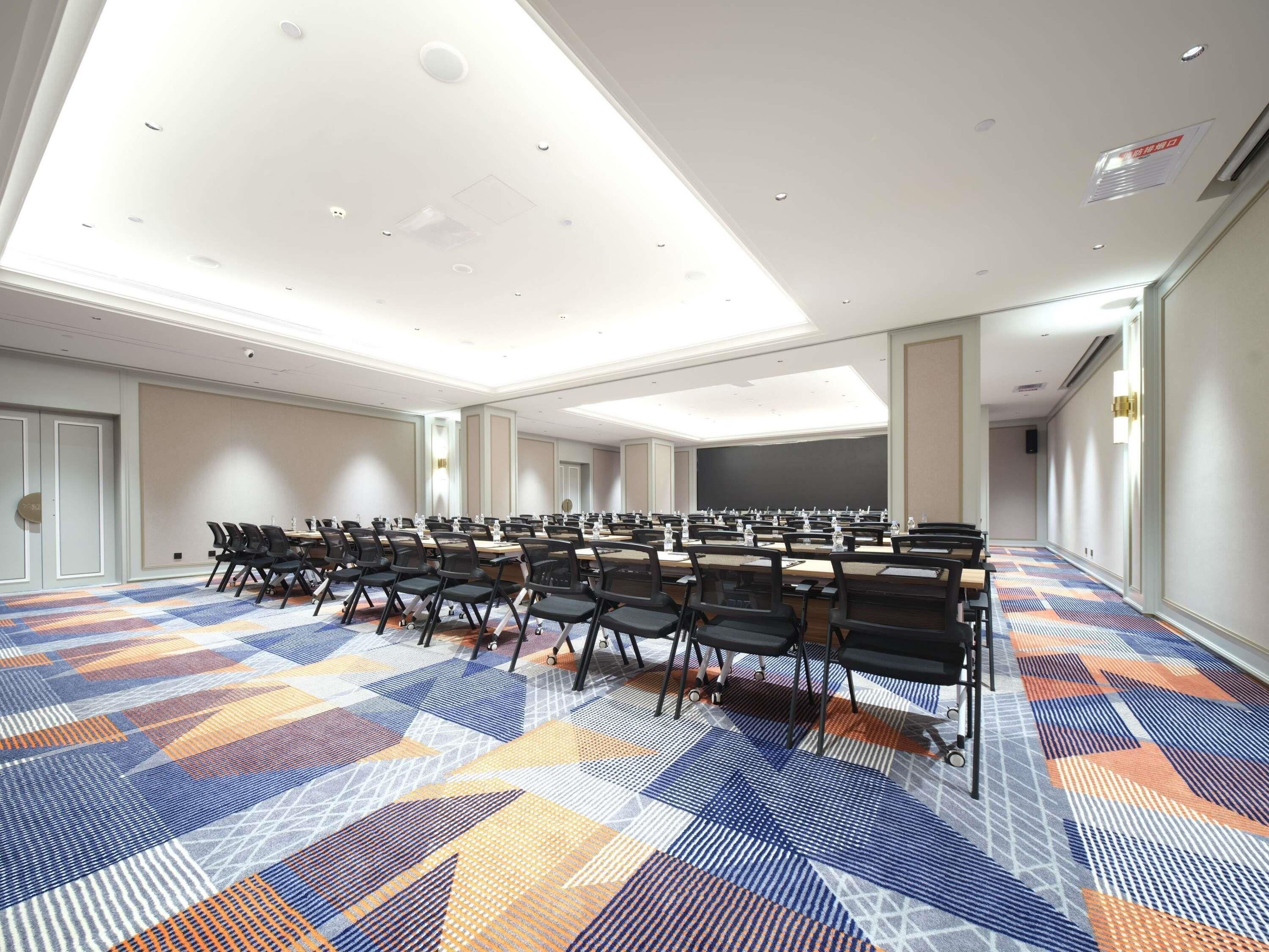 Sala de conferência com carpete estampado, mesas compridas e cadeiras, equipada com garrafas de água e papéis.