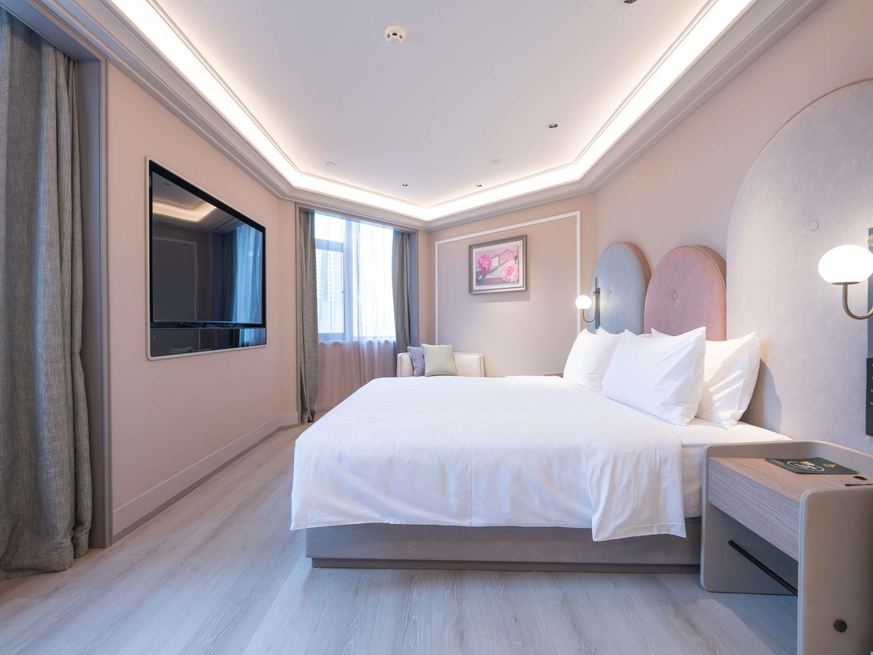 A modern hotel room with a large bed, a flat-screen TV, a bedside table, and a wall-mounted light fixture.