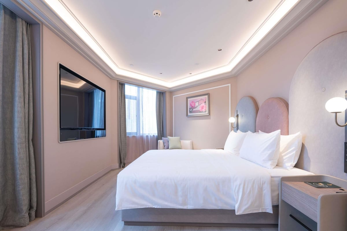 A modern hotel room with a large bed, a flat-screen TV, a bedside table, and a wall-mounted light fixture.