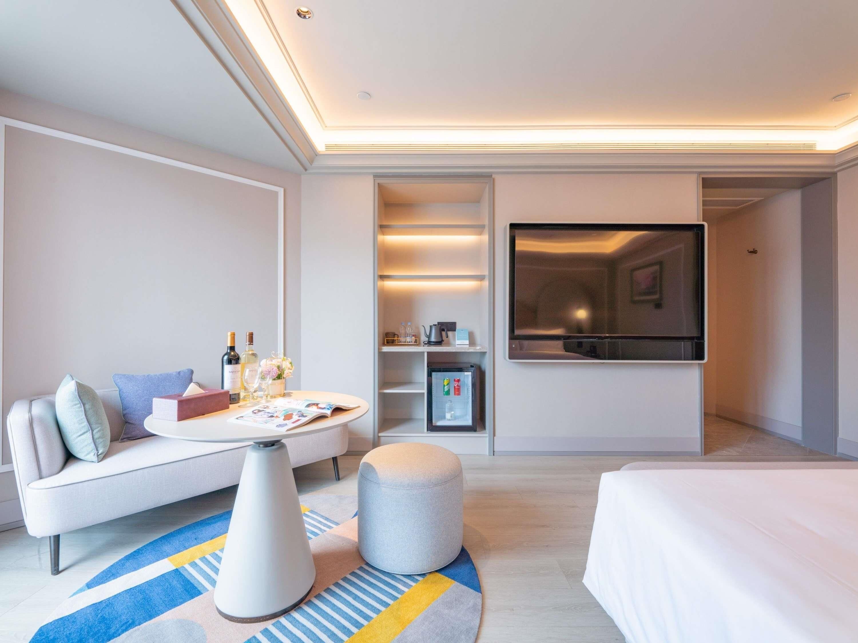 A modern hotel room with a sofa, ottoman, bedside table, and a large window.