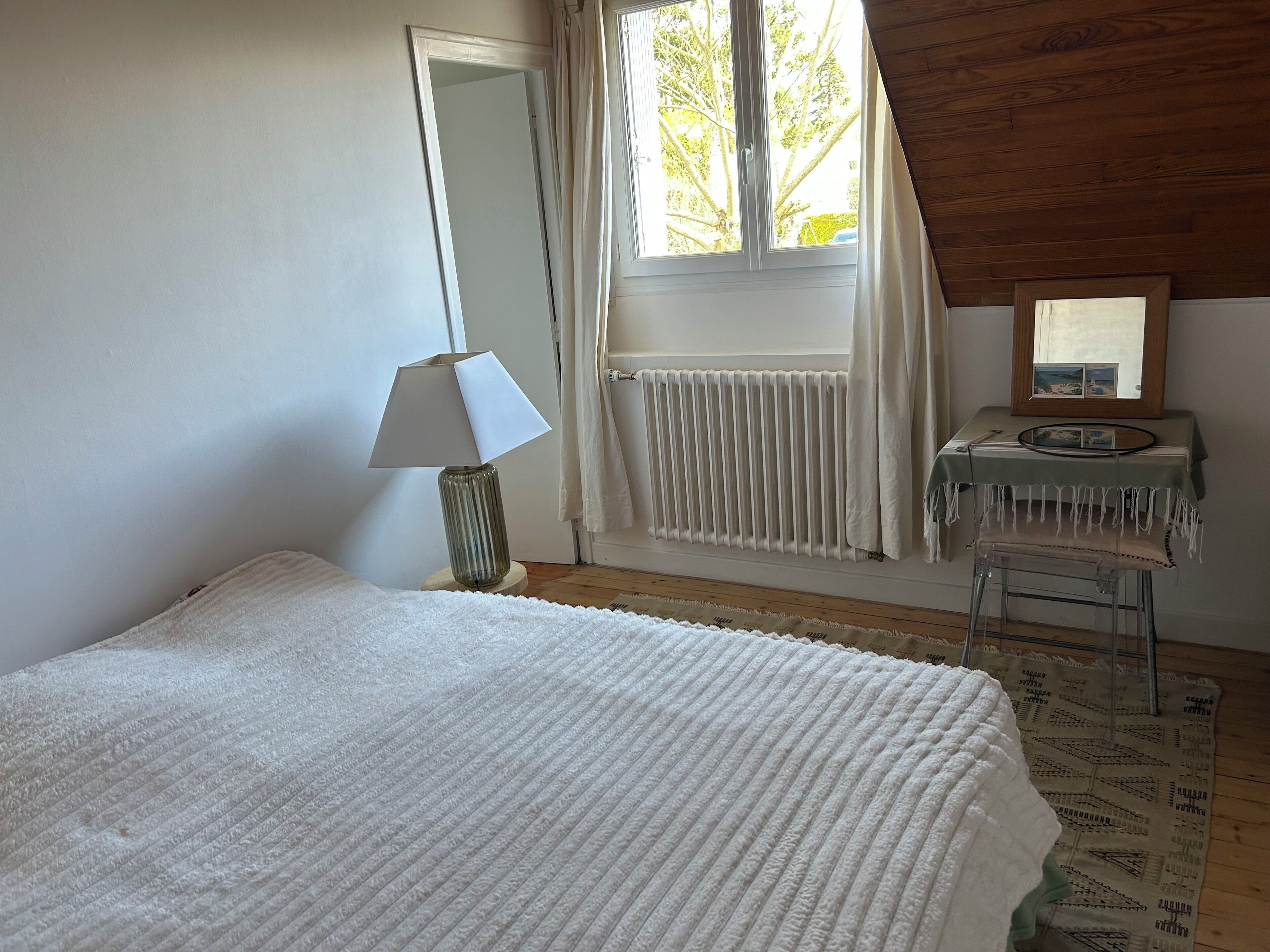 A bedroom with a bed, a lamp, a radiator, a window with curtains, a small table with a mirror, and a patterned rug.