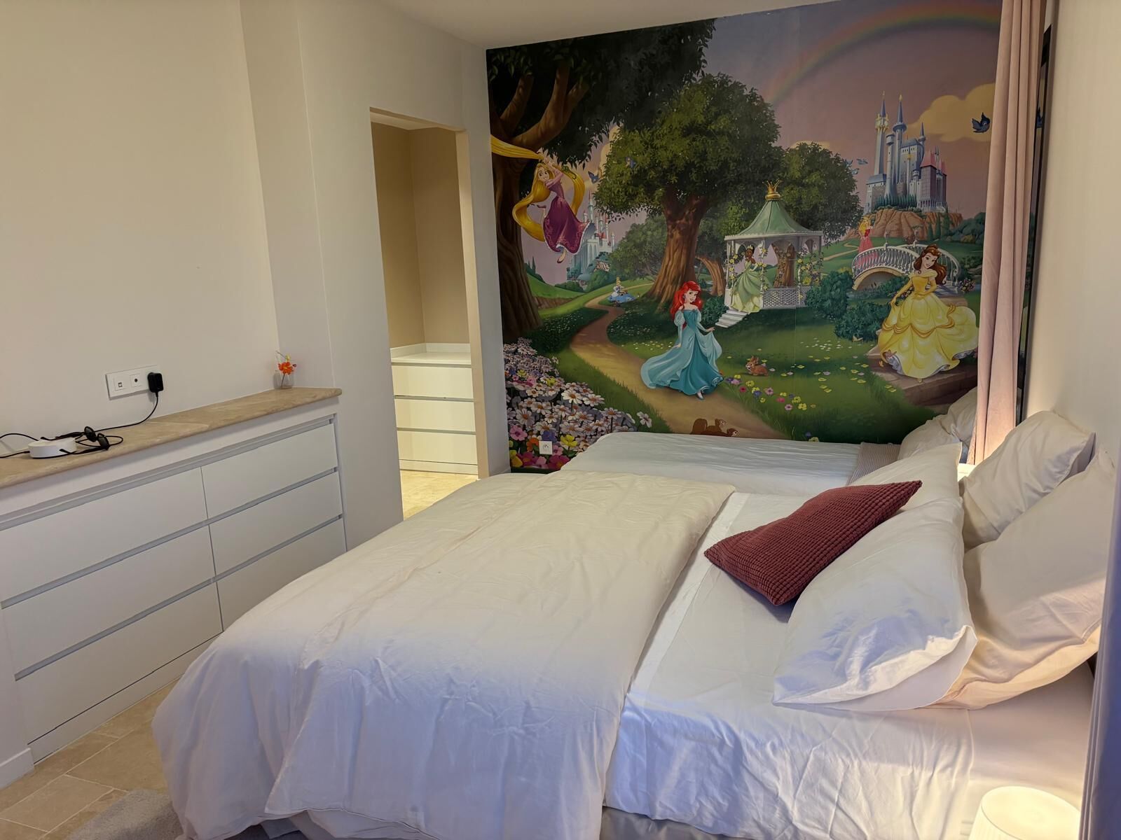 Une chambre avec une fresque représentant un château, un lit avec une literie blanche et une commode.