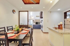 Uma sala de jantar com uma mesa comprida posta para refeição, uma cozinha com geladeira e bancada, e uma escada que leva a um andar superior.