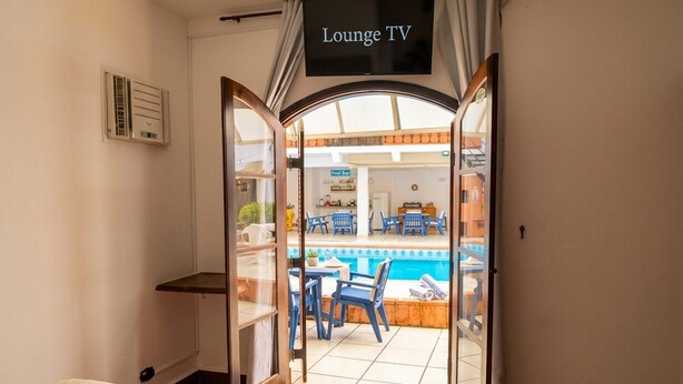Una habitación con cama, un televisor que dice 'Lounge TV' y vistas a un área de piscina exterior con sillas y mesas.