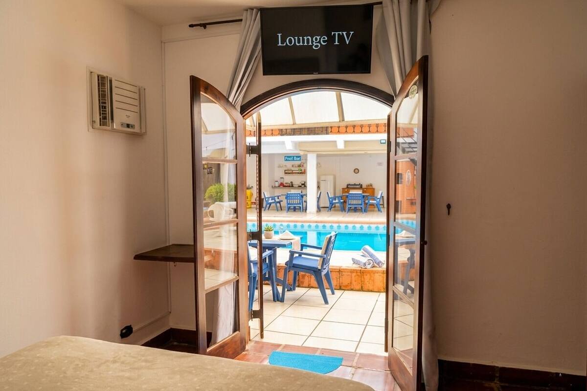Una habitación con cama, un televisor que dice 'Lounge TV' y vistas a un área de piscina exterior con sillas y mesas.