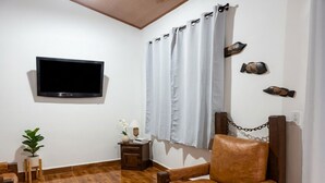 Habitación con televisión de pared, un sillón de cuero, una mesita con lámpara, una planta en maceta y cortina con cadena.
