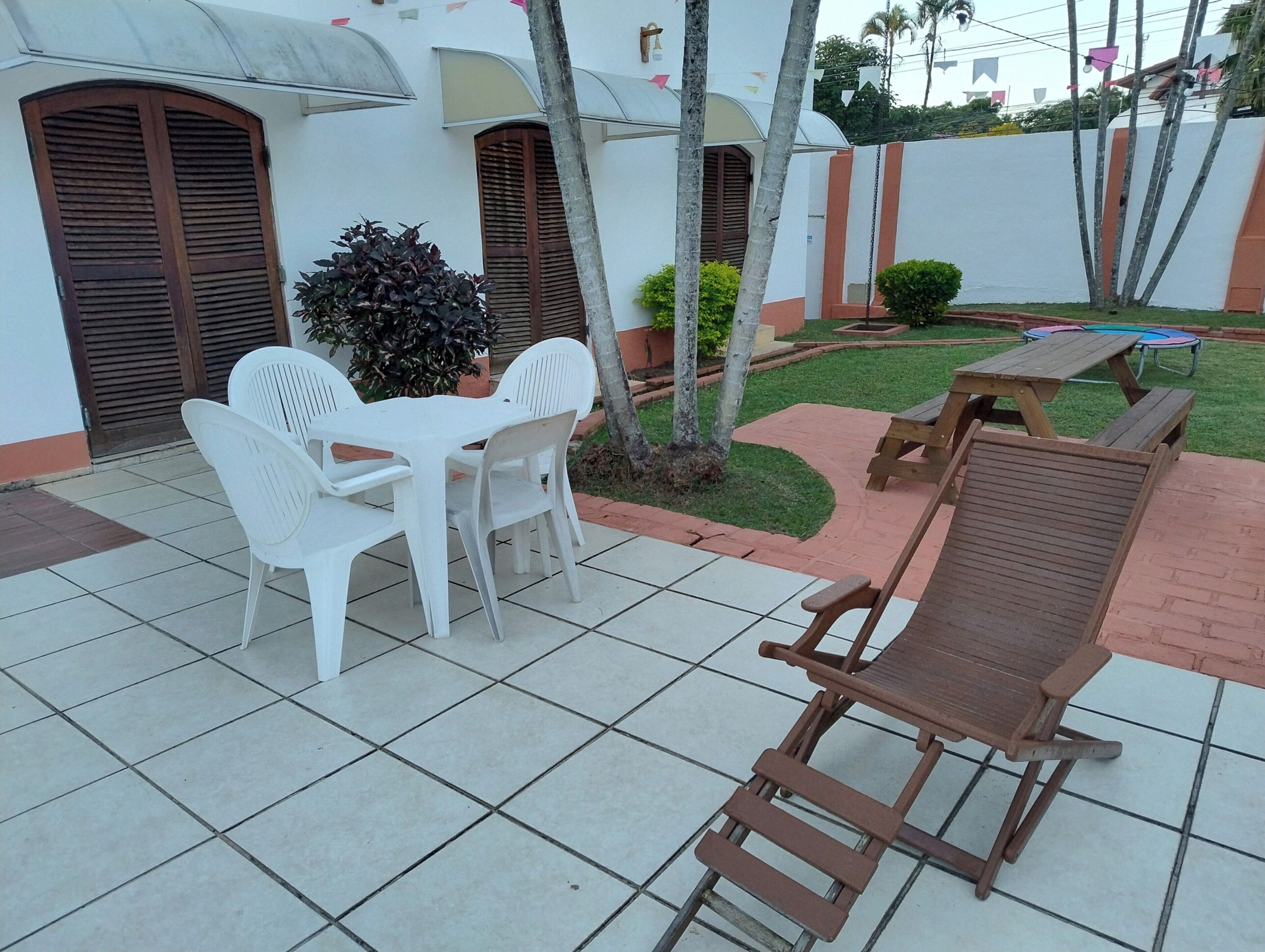 A patio with a table and chairs, a wooden lounge chair, and a small garden area with a tree and a bench.