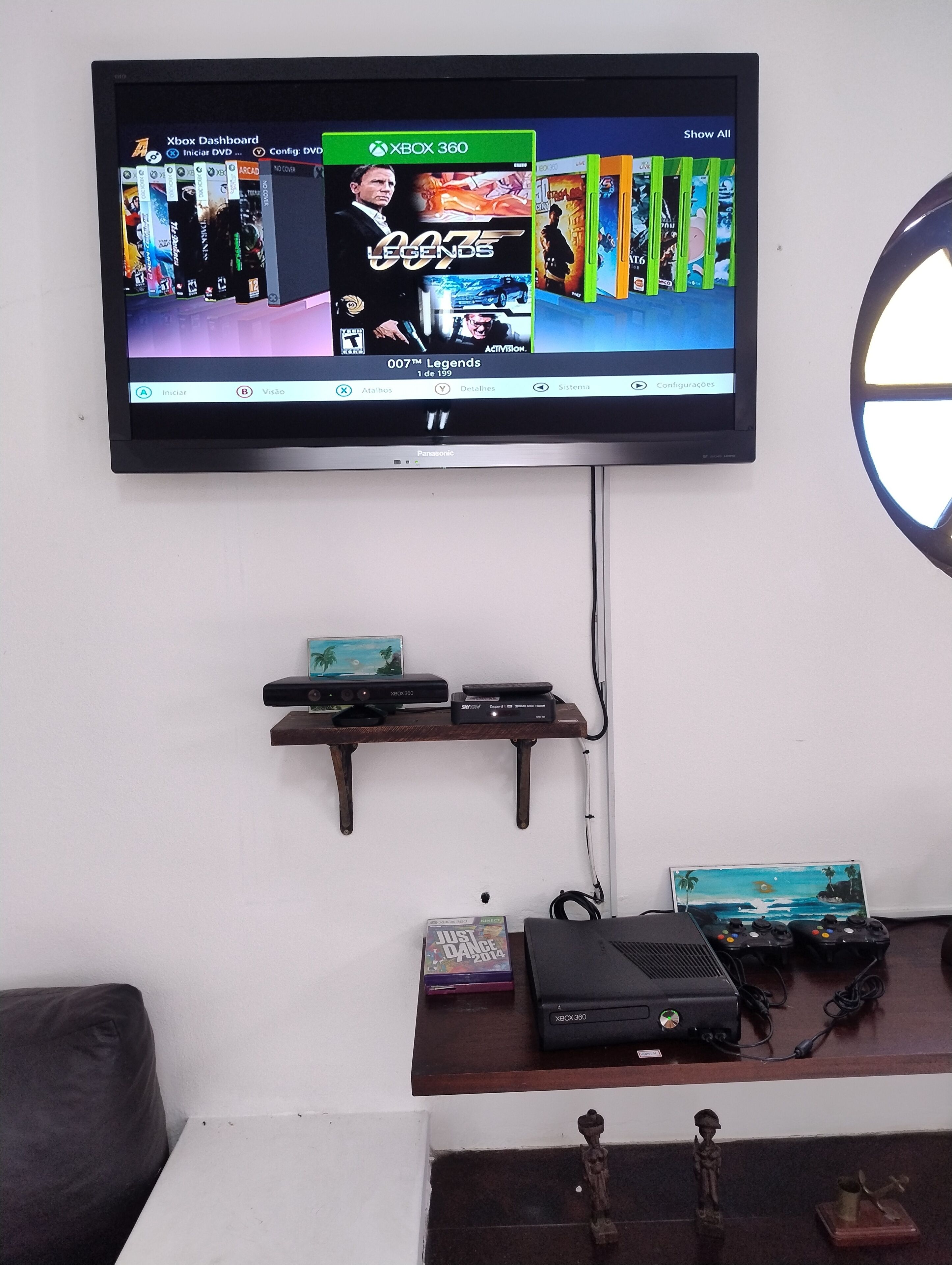 Un televisor montado en la pared muestra la interfaz de Xbox 360. Debajo del televisor, hay un estante de madera con una consola PlayStation y un reproductor de DVD.