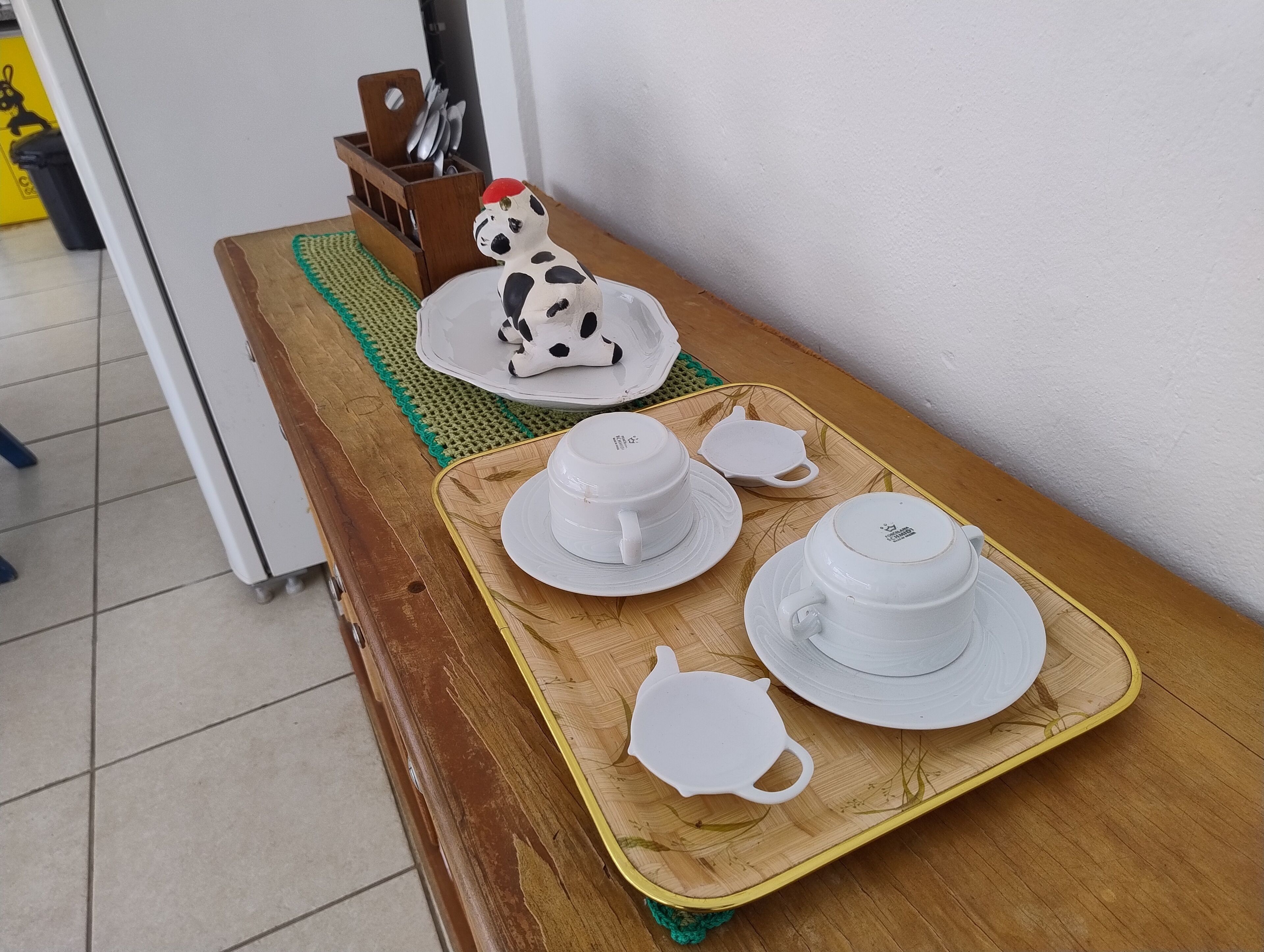 Una mesa de madera con una bandeja que sostiene tazas y platillos blancos, un calentador de té y una pequeña figurita de vaca.