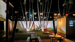 Un patio al aire libre con una mesa y sillas de madera, rodeado de palmeras y una pared blanca con una ventana.