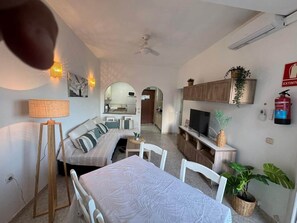 Ein modernes Wohnzimmer mit einer Couch, einem Esstisch und einem Fernseher. Im Hintergrund befindet sich eine Küchenzeile.