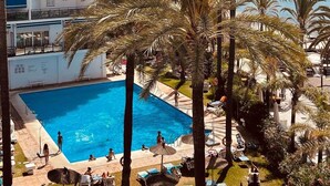 Ein mehrstöckiges Hotel mit Schwimmbad, umgeben von Palmen und Liegestühlen.