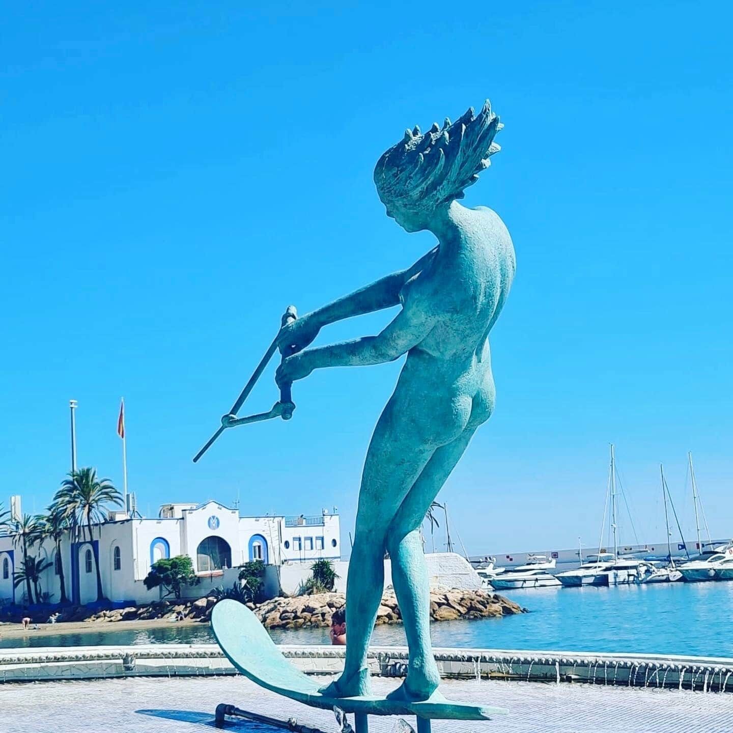 Eine Statue einer Figur, die ein Speer hält, befindet sich in der Nähe eines Uferbereichs mit Gebäuden und Booten im Hintergrund.