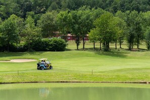 Campo de golf con un obstáculo acuático, árboles y un edificio al fondo.