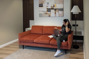 Una persona sentada en un sofá con una computadora portátil, una lámpara y un cuadro en la pared.