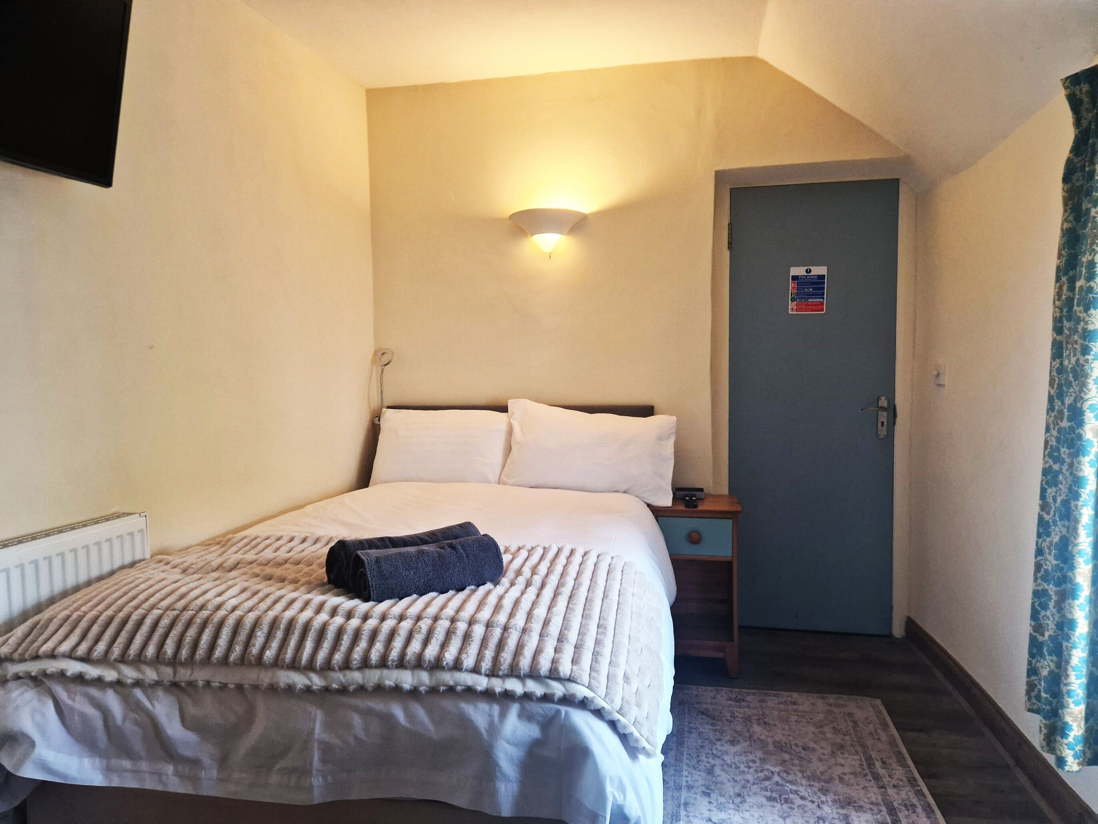 A hotel room with a bed, bedside table, lamp, and a door with a sign.