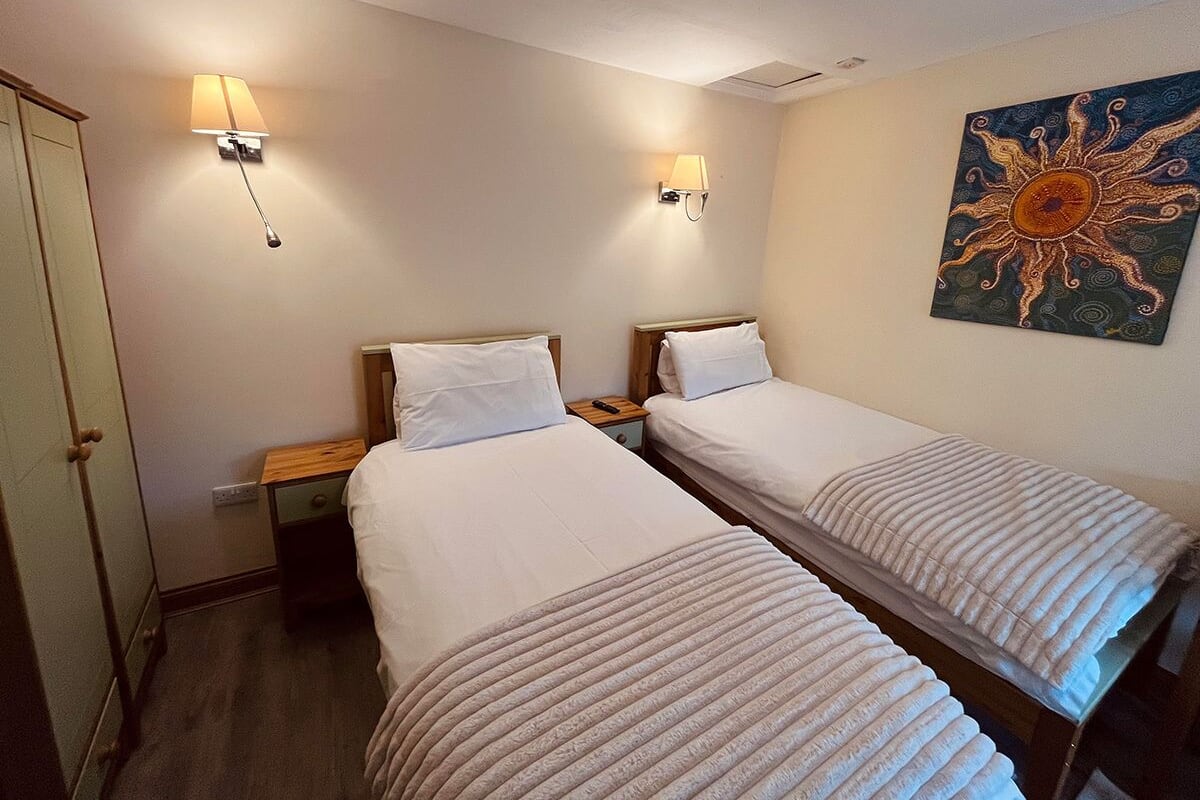 Camera d'albergo con due letti singoli, lampade da comodino, un comodino in legno, una porta e un quadro appeso al muro.