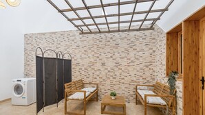 A room with a washing machine, two wooden benches, a small table with a plant, a black partition, and a brick wall.