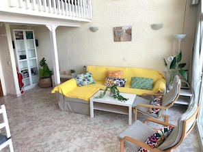 Ein gemütlicher Wohnbereich mit einem gelben Sofa, einem weißen Couchtisch und zwei Holzstühlen. Ein weißes Schränkchen und ein Blumentopf sind ebenfalls zu sehen.