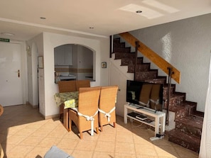 A living room with a TV, wicker chairs, a staircase, and a kitchen area. - 1.Skol Marbella 703A (Marbella)