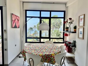 A dining area with a round table, white chairs, a large window with a view of palm trees and the sea, a pendant light, and a shelf with potted plants. - 2. Skol Marbella 337B (Marbella)