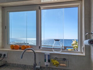 Eine Küche mit Fenster zum Meer, einem Wasserhahn, einem Glasbehälter und einem Geschirrgestell mit einem gelben Handtuch.