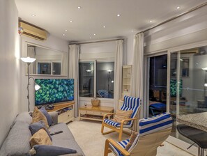 Ein Wohnzimmer mit einer Couch, Sesseln, einem Fernseher und einer Glastür, die auf einen Balkon führt.