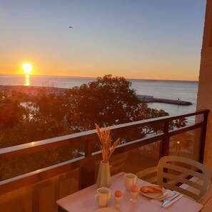Ein Tisch mit einer Vase, Tassen und einem Teller Essen auf einem Balkon mit Blick auf den Sonnenuntergang über dem Meer.