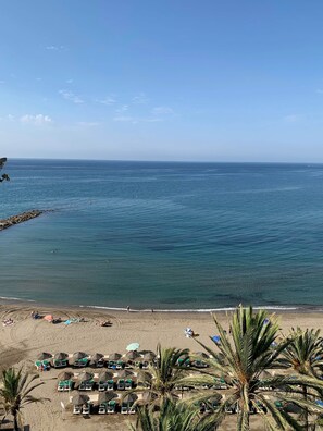 Ein Strand mit Liegestühlen und Sonnenschirmen, blaues Meer und ein Felsenufer.