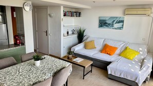 Una sala moderna con comedor, un sofá blanco con cojines de colores, una mesa de centro y una estantería.