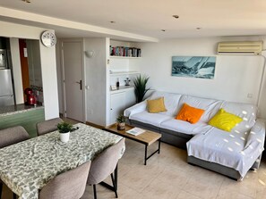 Ein modernes Wohnzimmer mit Essbereich, einem weißen Sofa mit bunten Kissen, einem Couchtisch und einem Bücherregal.