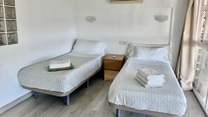 Habitación de hotel con dos camas individuales, una mesita de noche de madera, aire acondicionado instalado en la pared y una ventana con cortinas.