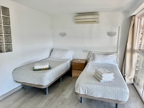 Ein Hotelzimmer mit zwei Einzelbetten, einem kleinen Nachttisch aus Holz, einer Wandklimaanlage und einem Fenster mit Vorhängen.