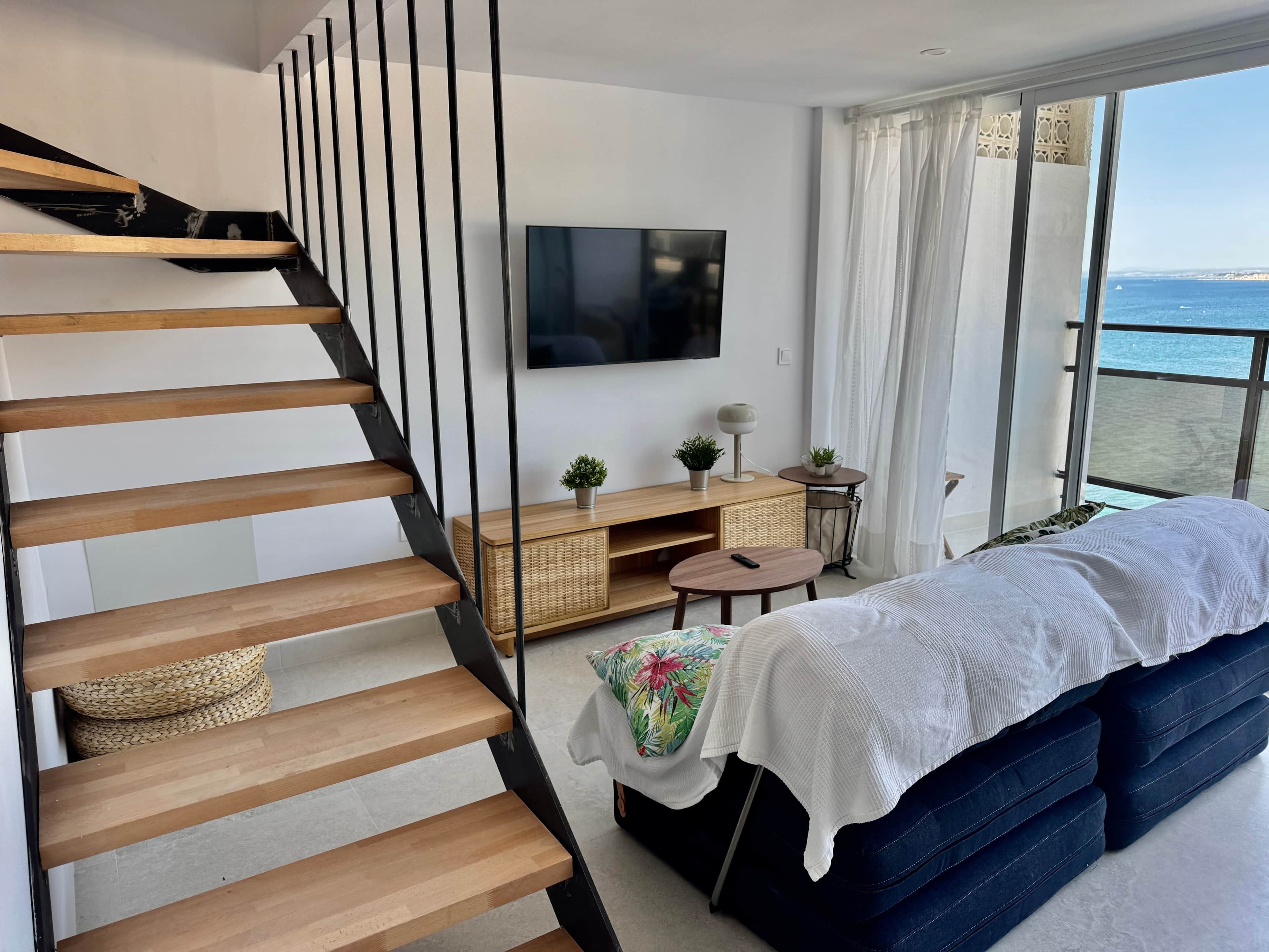 A modern bedroom with a wooden staircase, a bed with a white cover, a TV, a small table, and a view of the sea.