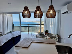 Ein Schlafzimmer mit einem Bett, einem Nachttisch, einem kleinen Tisch mit Pflanze und Blick aufs Meer.