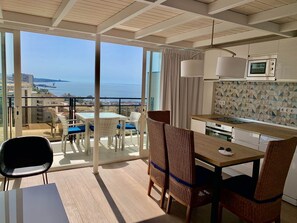 Eine moderne Küche mit weißen Schränken und einer geometrischen Spritzschutzwand, ein Essbereich mit Korbstühlen und einem Glastisch sowie ein Balkon mit Meerblick.
