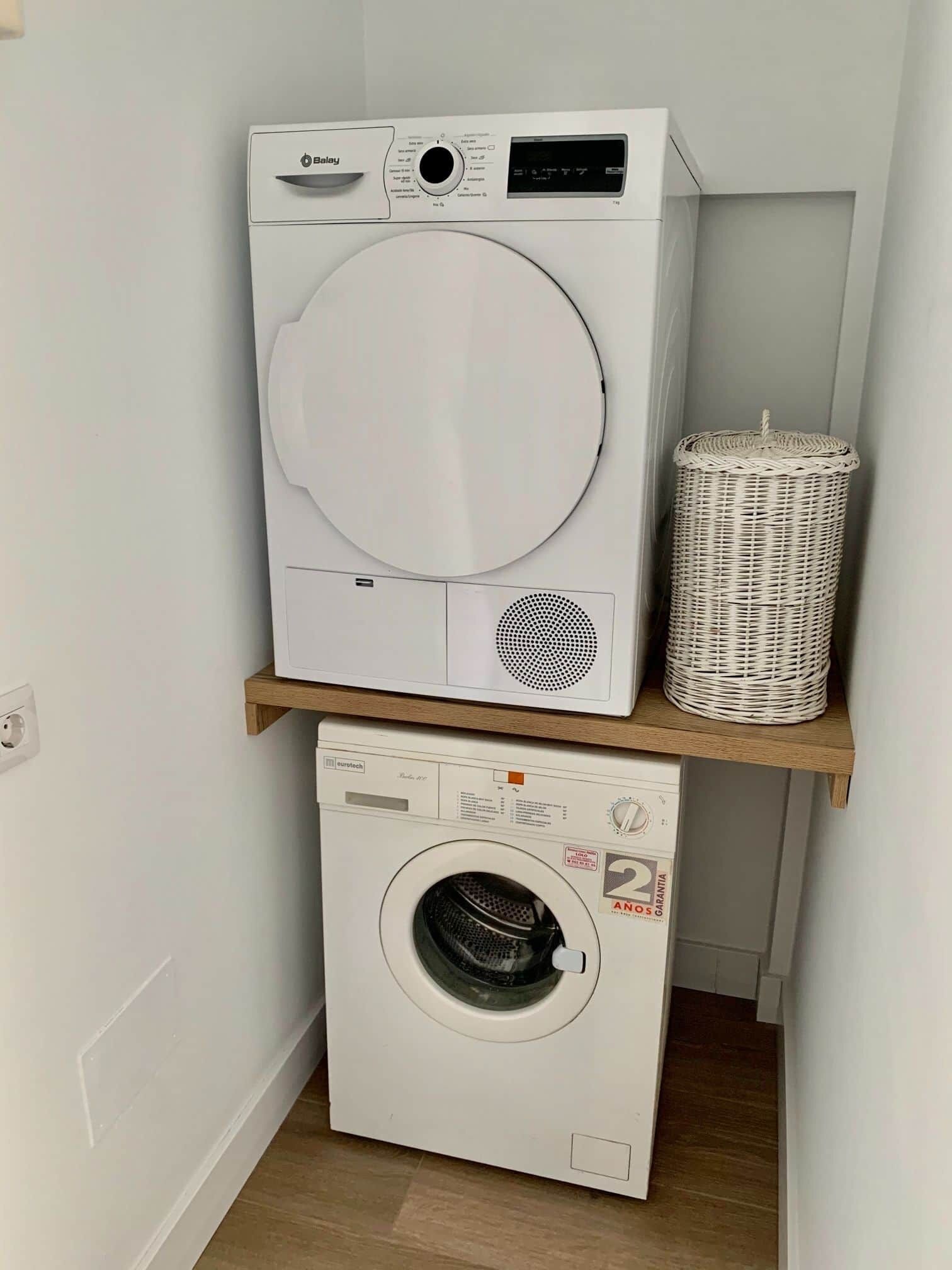 Ein Waschraum mit einer Haube Waschemaschine und einem Trockner, die übereinander auf einem Holzregal stehen, einem Korbwäscheschrank auf dem Regal und einer Steckdose an der Wand.