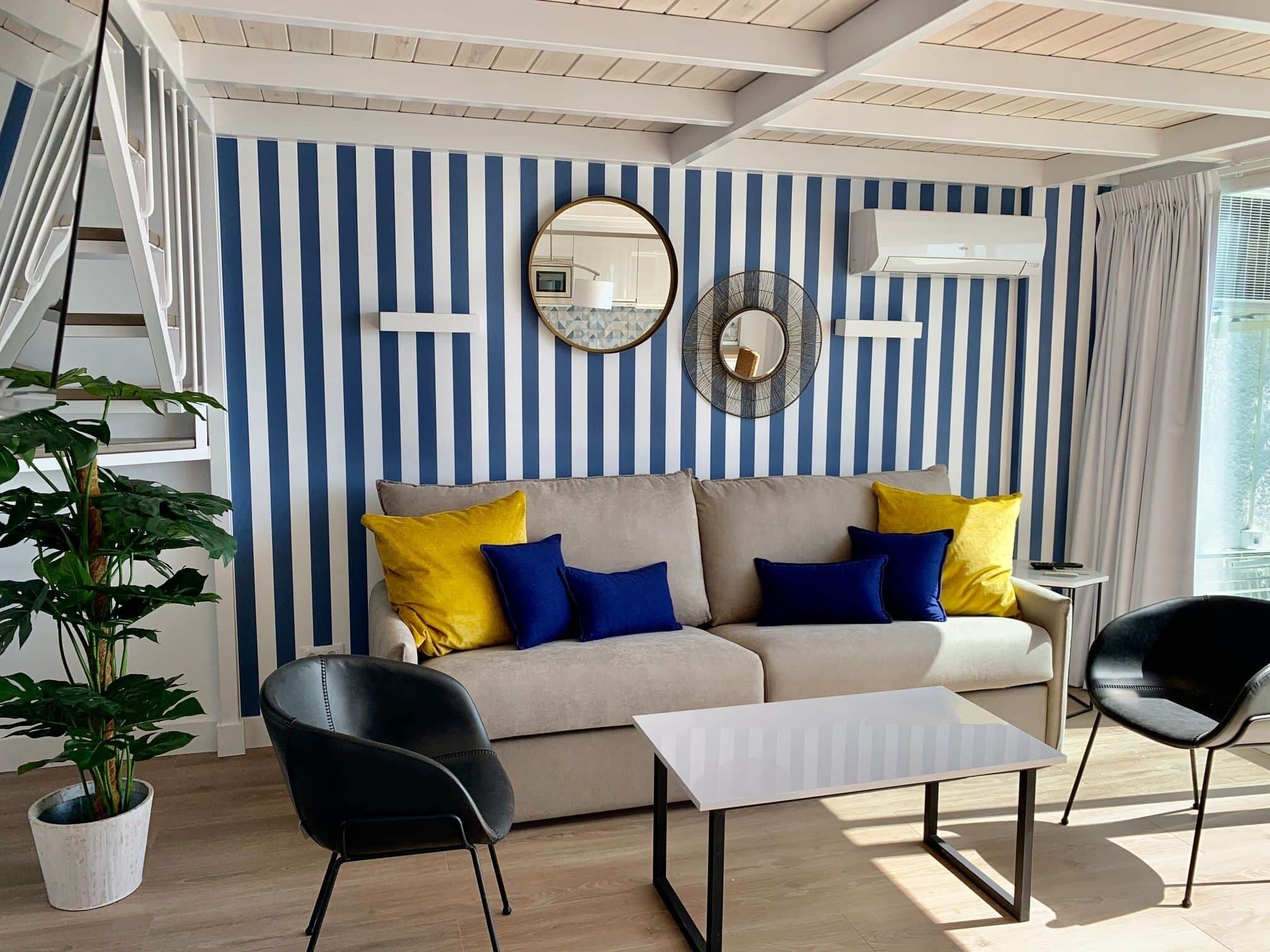Ein modernes Wohnzimmer mit einer gestreiften Wand, einer Couch mit Kissen, einem Couchtisch, zwei Sesseln, einer Topfpflanze und einer Klimaanlage.