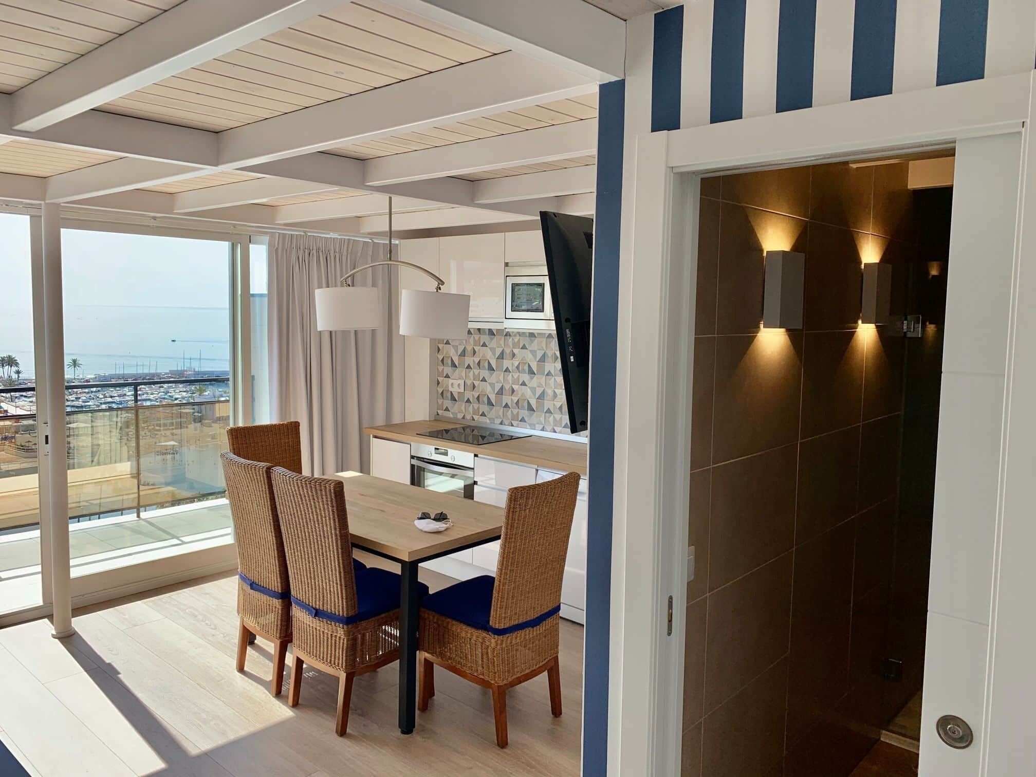 A modern room with a dining table, chairs, a kitchen area, and a balcony with a view.