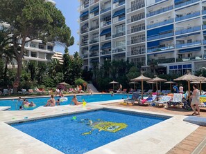 Una piscina con sillas de descanso y sombrillas, rodeada de vegetación y un edificio de varios pisos.