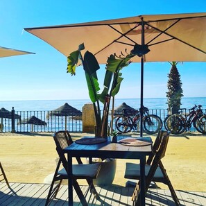 Ein Tisch mit Stühlen und ein großer Sonnenschirm mit Blick auf einen Strand, wo Fahrräder und ein Zaun stehen.
