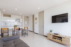 A modern living room with a sofa, a TV mounted on the wall, a wooden cabinet with drawers, and a kitchen area in the background. - U-GO Ocean Club Confort Bello Horizonte (Santa Marta)