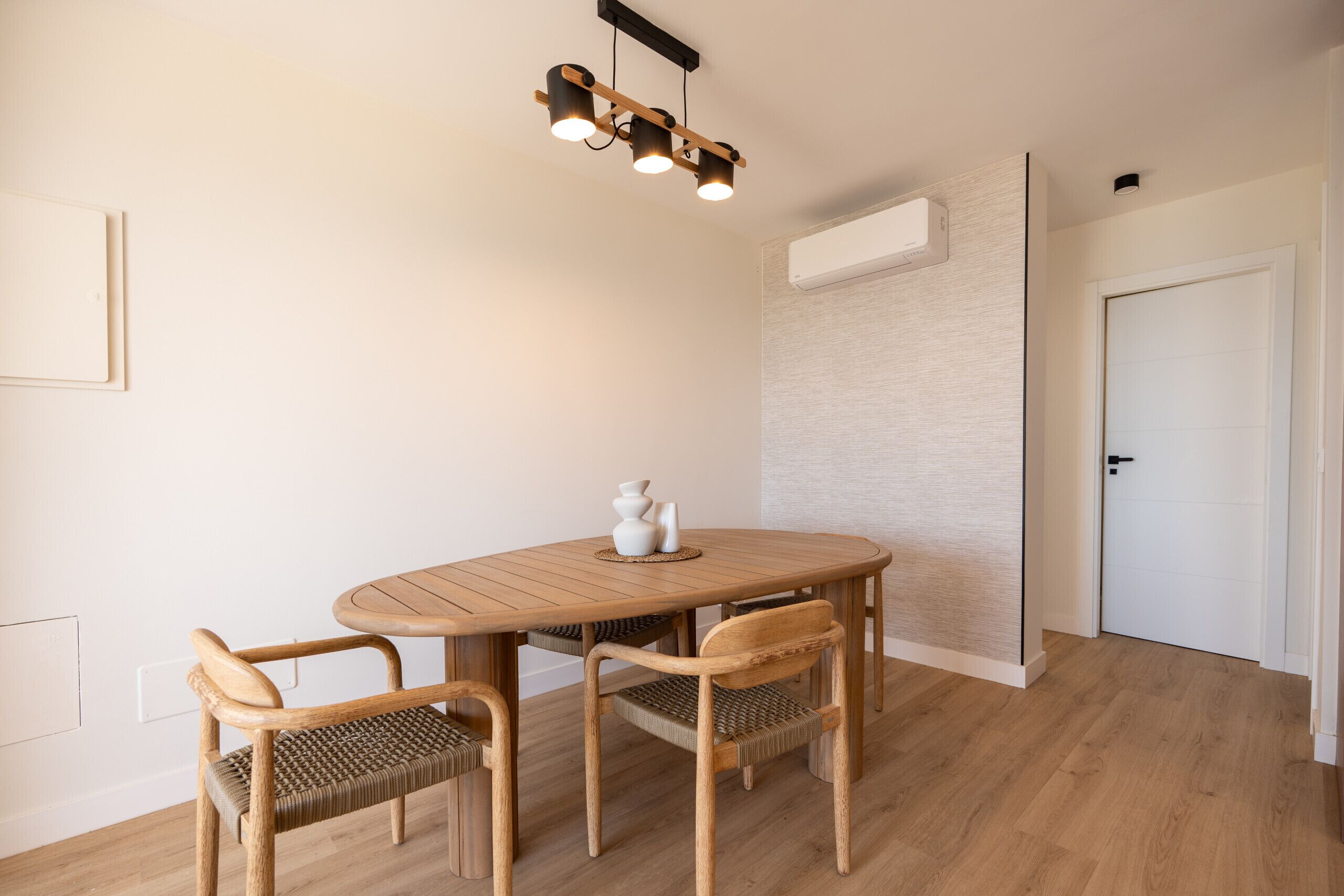 Un coin repas avec une table ronde en bois, quatre chaises, un luminaire suspendu moderne et une porte blanche.