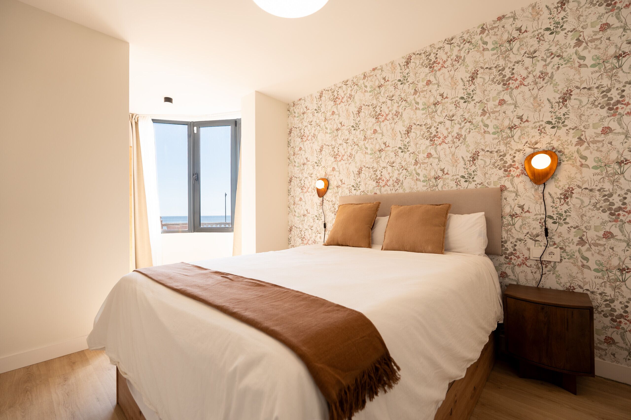 Un dormitorio con una cama, una mesita de noche, una lámpara de pared y una ventana con cortinas.